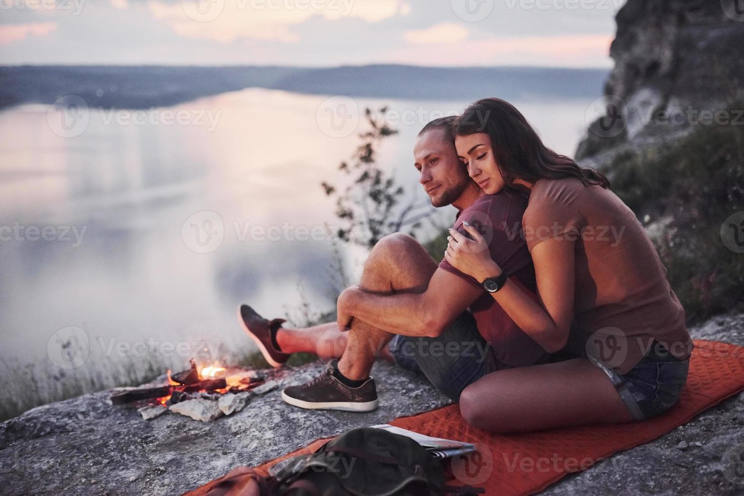 casal abraçando com mochila sentado perto do fogo no topo da montanha, apreciando a vista da costa de um rio ou lago. viajando ao longo das montanhas e da costa, conceito de liberdade e estilo de vida ativo foto