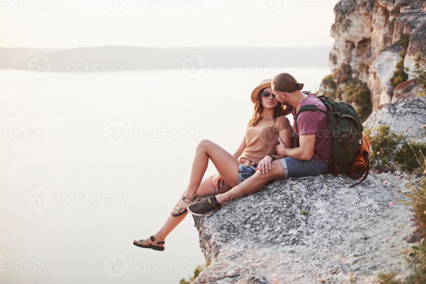 casal abraçando com mochila sentado no topo da montanha rochosa, apreciando a vista da costa de um rio ou lago. viajando ao longo das montanhas e da costa, conceito de liberdade e estilo de vida ativo foto