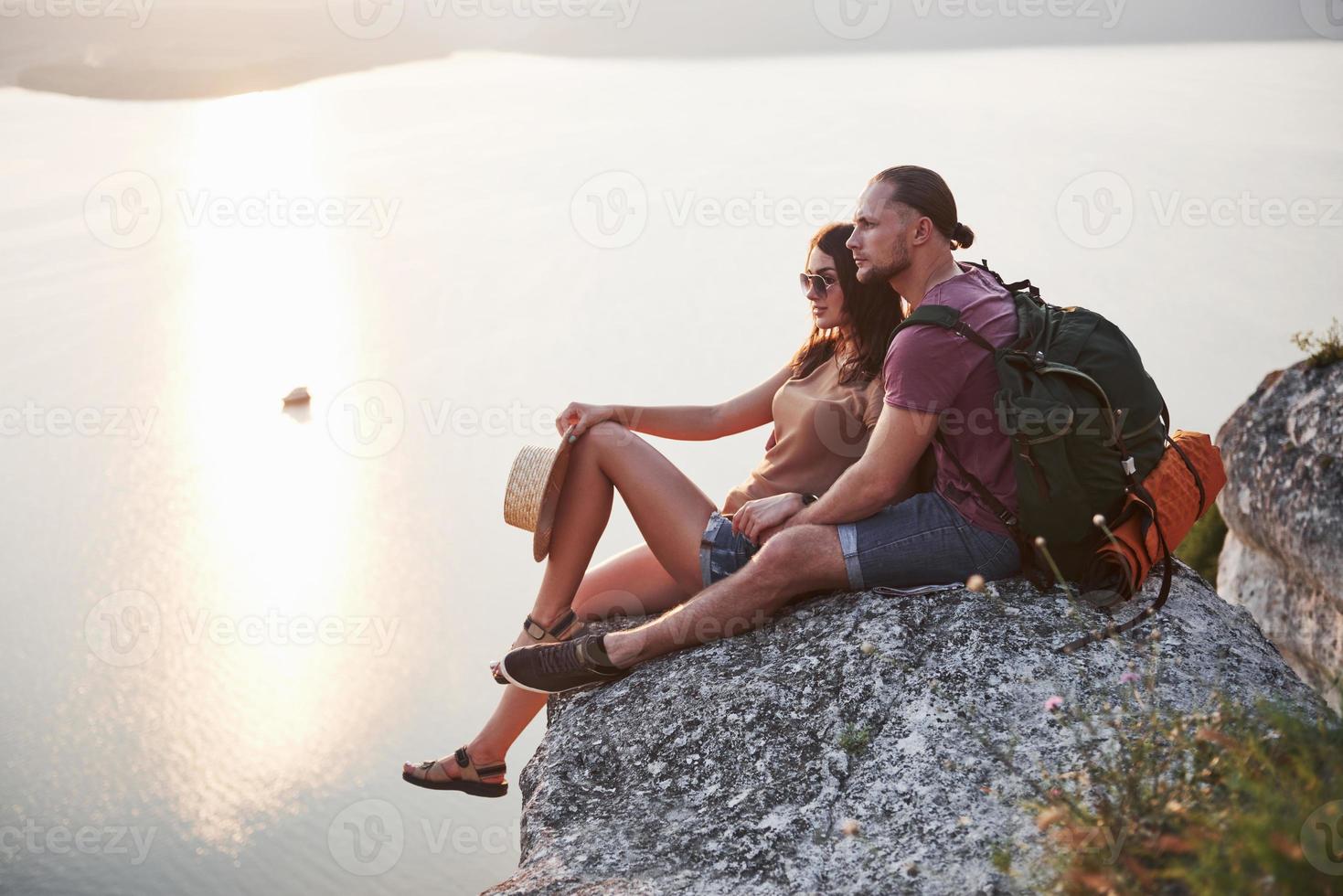 casal abraçando com mochila sentado no topo da montanha rochosa, apreciando a vista da costa de um rio ou lago. viajando ao longo das montanhas e da costa, conceito de liberdade e estilo de vida ativo foto