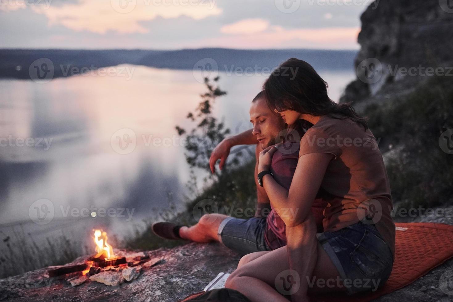 casal abraçando com mochila sentado perto do fogo no topo da montanha, apreciando a vista da costa de um rio ou lago. viajando ao longo das montanhas e da costa, conceito de liberdade e estilo de vida ativo foto