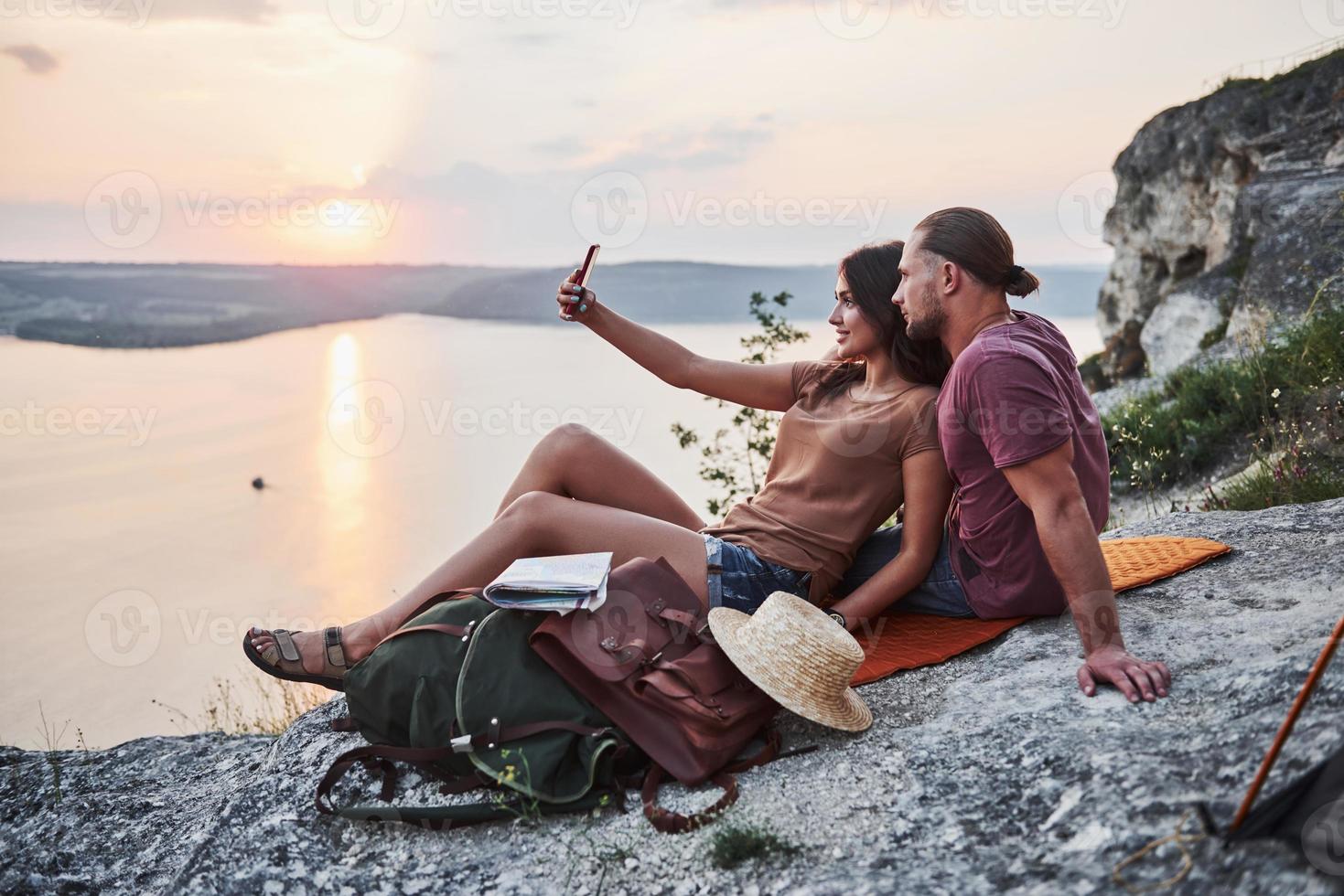 casal abraçando com mochila sentado no topo da montanha rochosa, apreciando a vista da costa de um rio ou lago. viajando ao longo das montanhas e da costa, conceito de liberdade e estilo de vida ativo foto