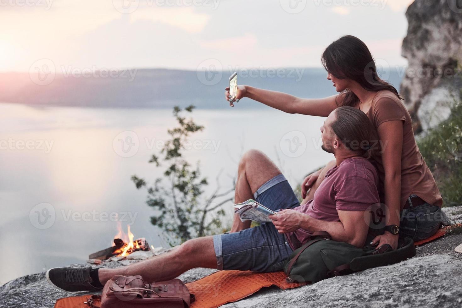 casal abraçando com mochila sentado perto do fogo no topo da montanha, apreciando a vista da costa de um rio ou lago. viajando ao longo das montanhas e da costa, conceito de liberdade e estilo de vida ativo foto