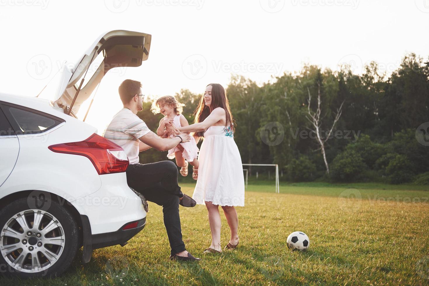 muito jovem casal e sua filha estão descansando na natureza. a mãe, o pai e a filha estão sentados no porta-malas do carro aberto foto