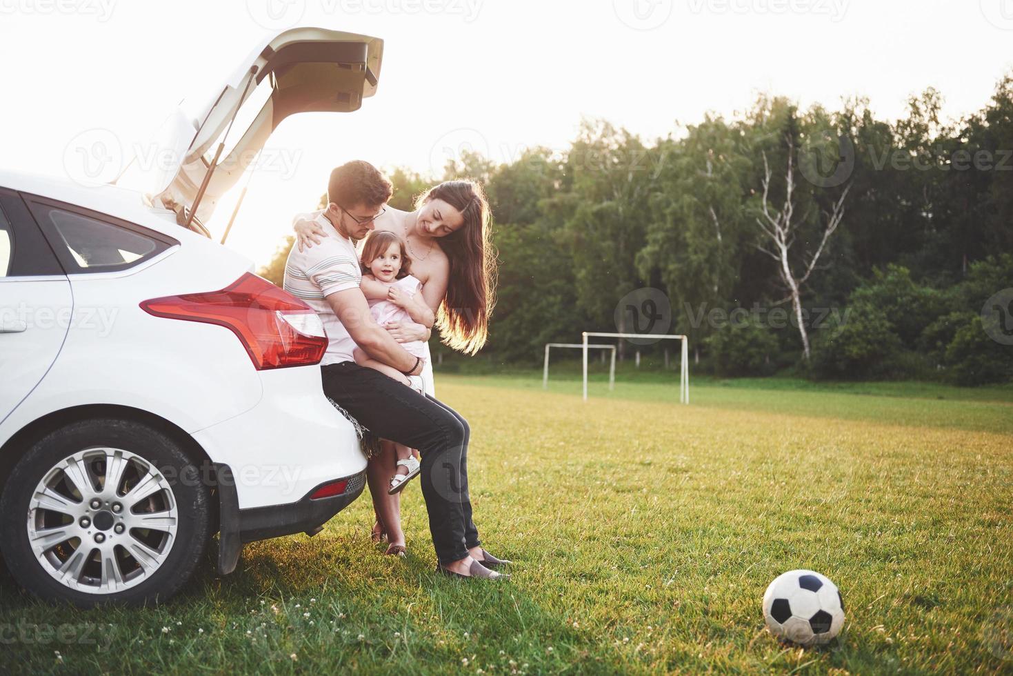 muito jovem casal e sua filha estão descansando na natureza. a mãe, o pai e a filha estão sentados no porta-malas do carro aberto foto