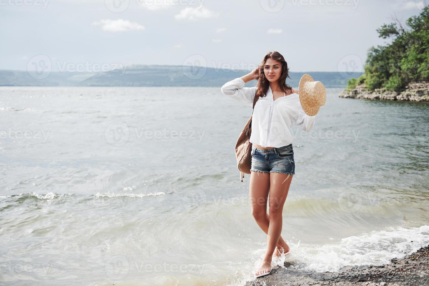 uma linda jovem com um chapéu e uma mochila caminha divertidamente à beira da água. um dia quente de verão é uma ótima época para aventura e aventura na natureza foto