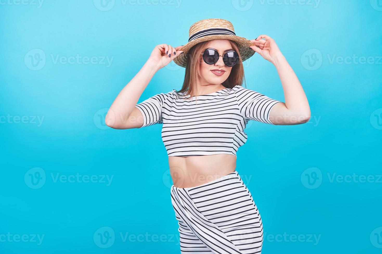 garota atraente em listras brancas e pretas, chapéu, óculos escuros, boca emocionalmente aberta sobre um fundo azul brilhante com um corpo perfeito. isolado. tiro de estúdio foto