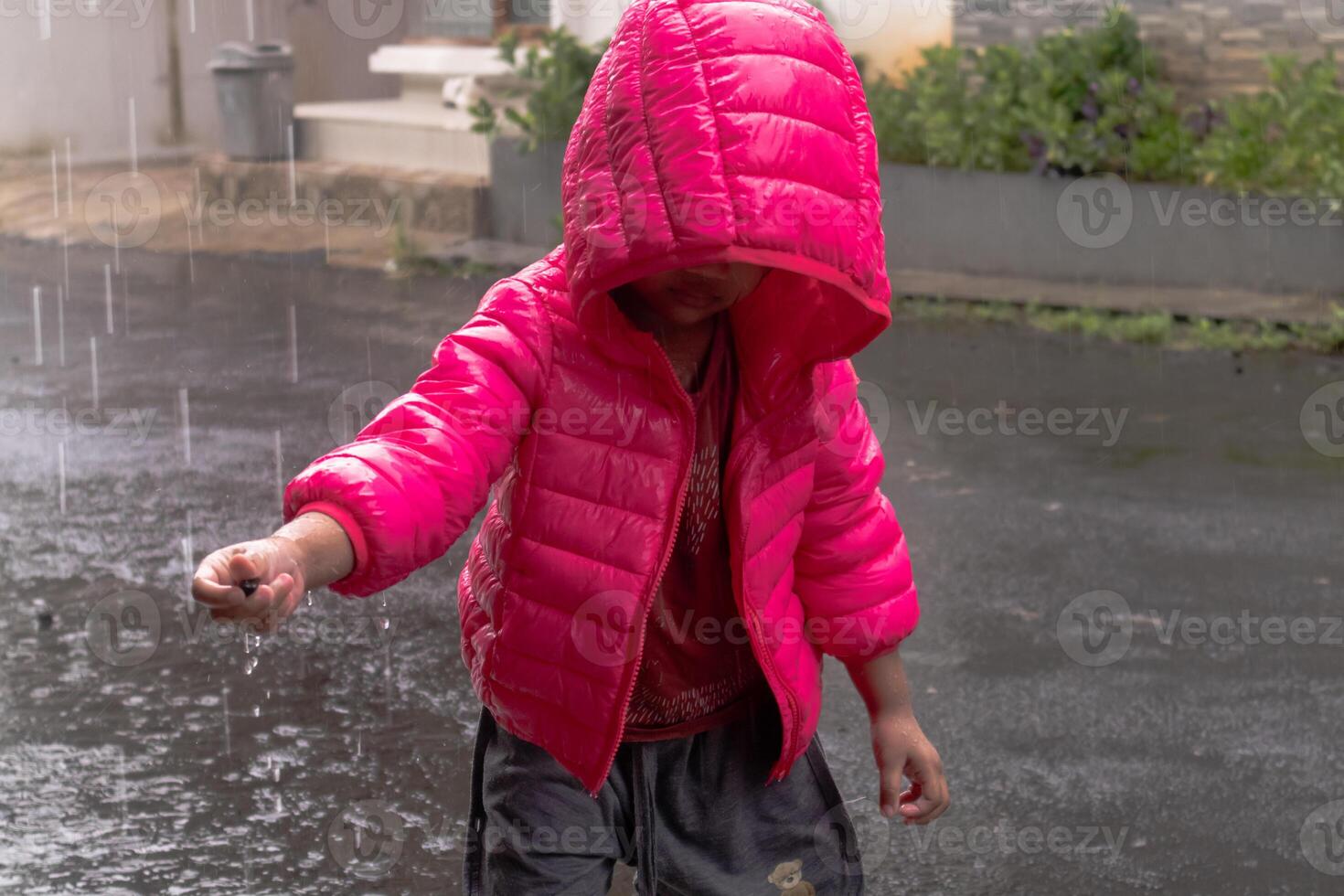 a ásia pequeno menina vestindo uma Rosa Jaqueta e jogando dentro a chuva dentro frente do a casa foto