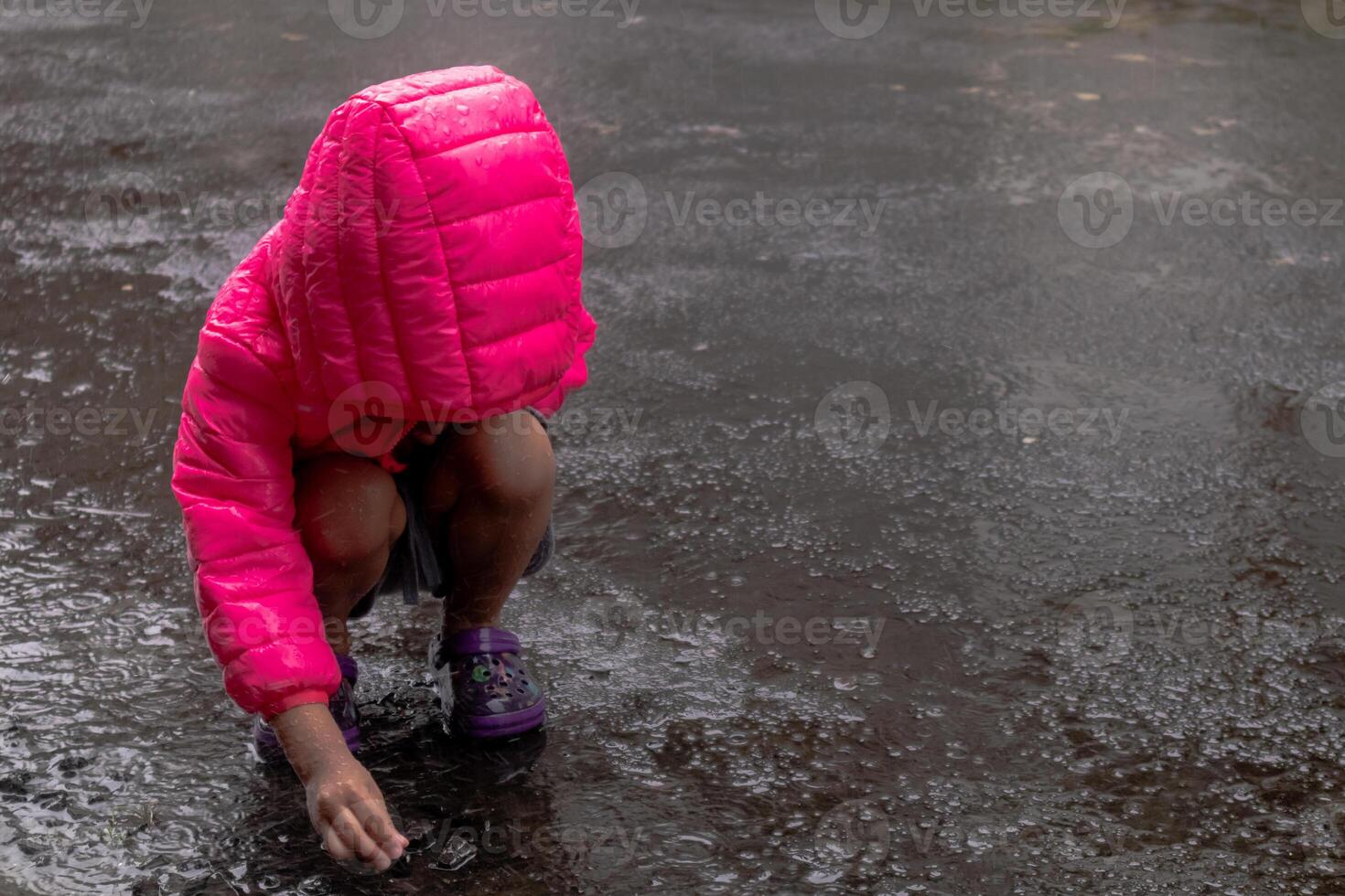 a ásia pequeno menina vestindo uma Rosa Jaqueta e jogando dentro a chuva em a asfalto estrada foto