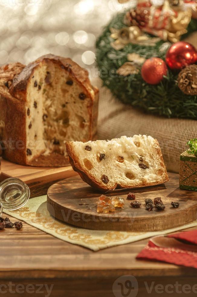 uma fatia de panetone e cubos de frutas cristalizadas em uma tábua de madeira com enfeites de natal foto