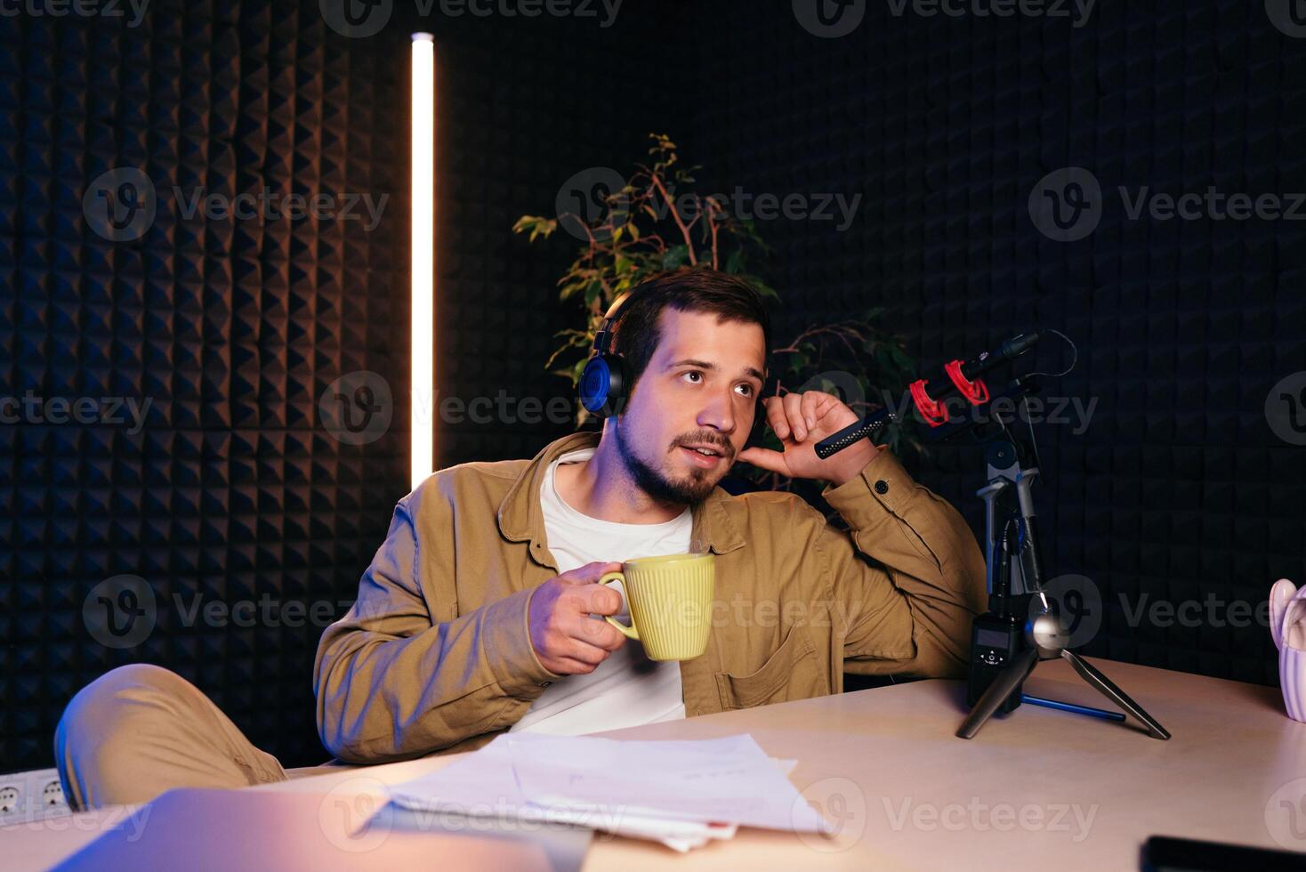 jovem à moda homem dentro mostarda camisa com fones de ouvido gesticulando às microfone e partilha história com público enquanto sentado às escrivaninha dentro estúdio com néon iluminação e gravação podcast foto