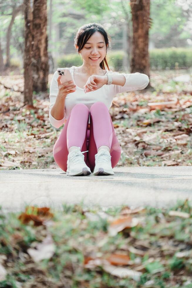 mulher sentado em uma parque caminho parece às dela ginástica rastreador enquanto segurando uma Smartphone, com espalhados outono folhas dentro a fundo. foto