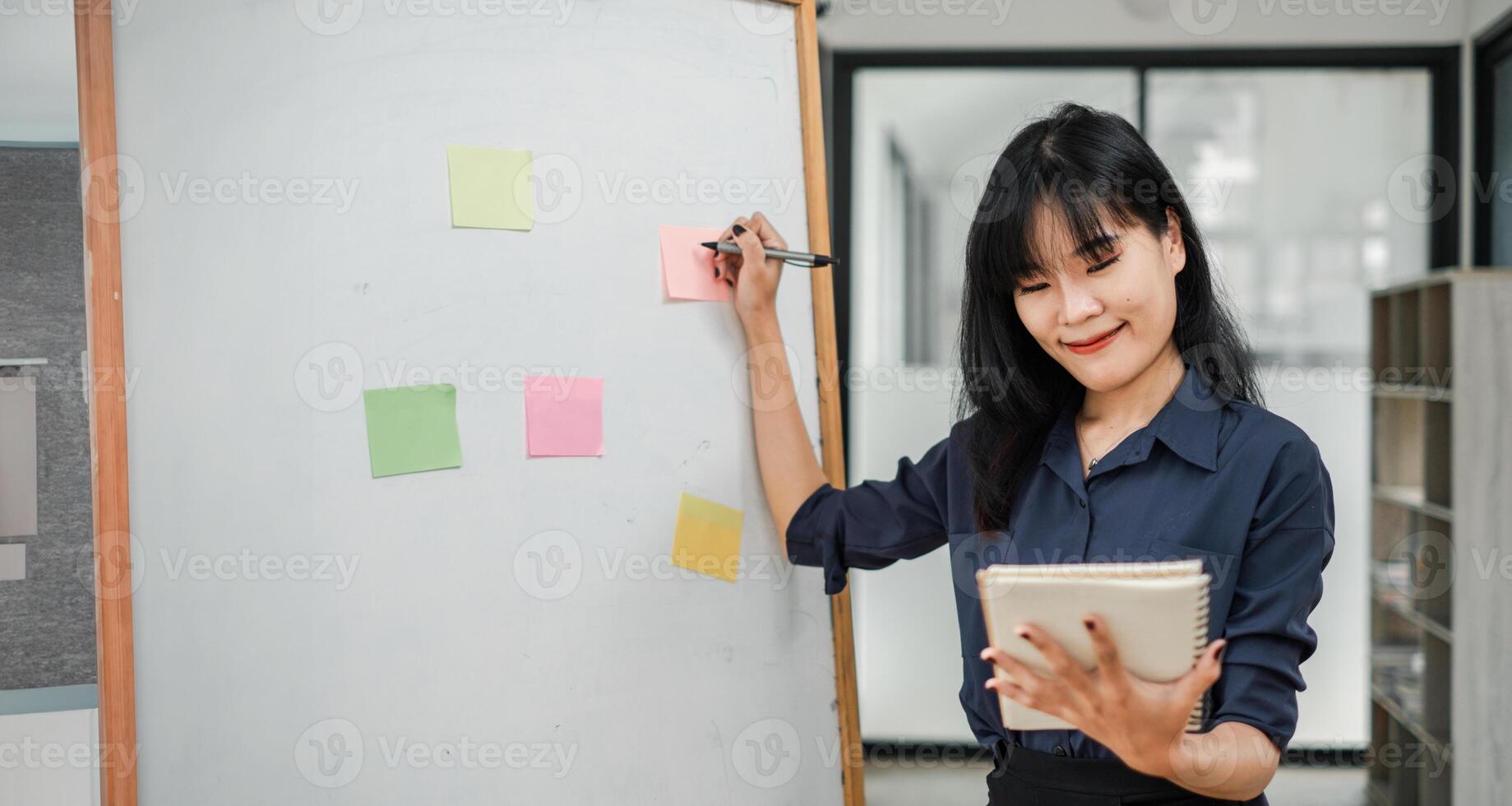 profissional mulher dentro a escritório configuração usando pegajoso notas em uma quadro branco para organização enquanto segurando uma planejador. foto