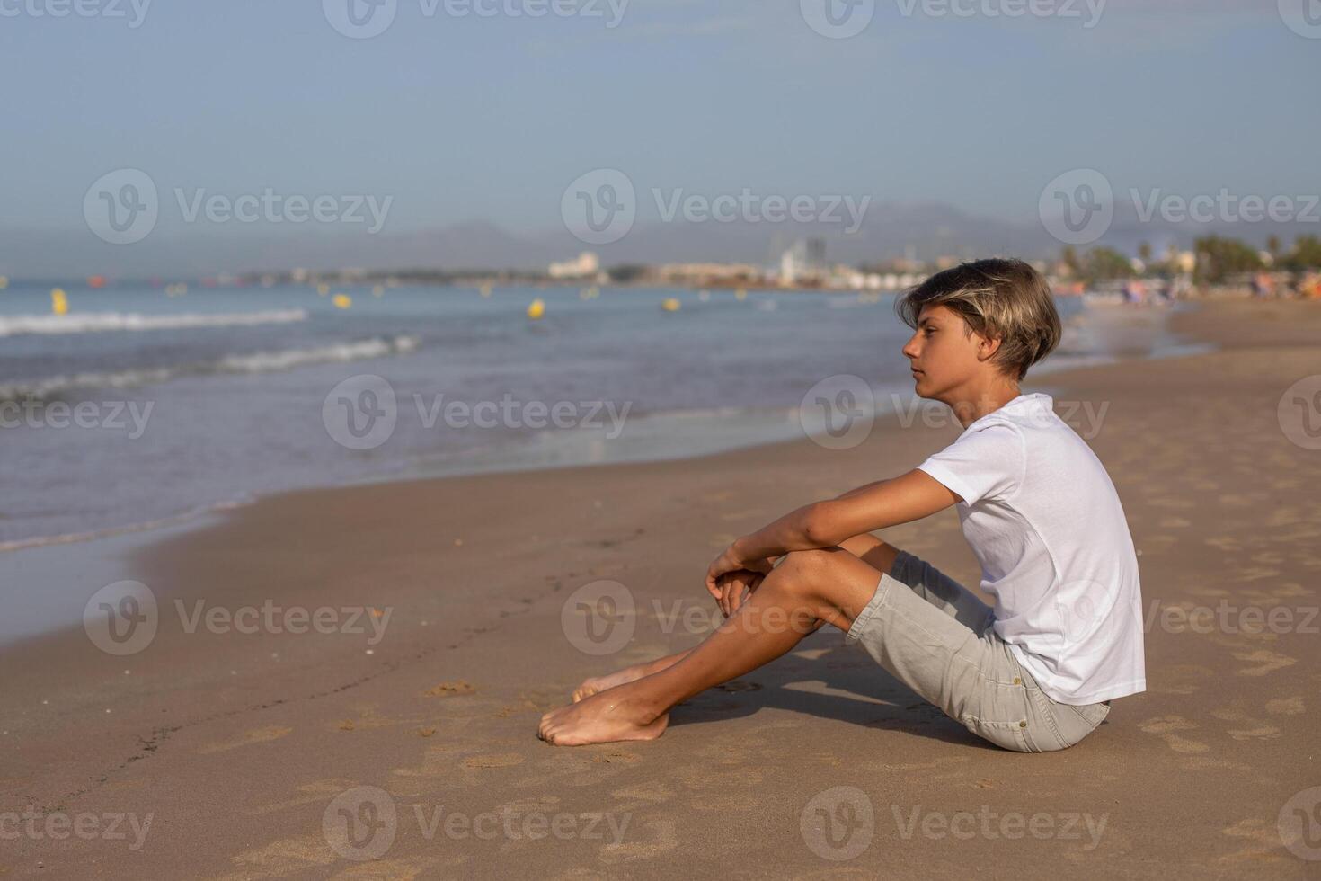 bonito adolescente Garoto dentro branco camiseta sentado às a de praia e apreciar uma verão período de férias olhando longe, salou cidade espanha.família feriados, férias, viagem, viagens conceito.side visualizar, cópia de espaço. foto
