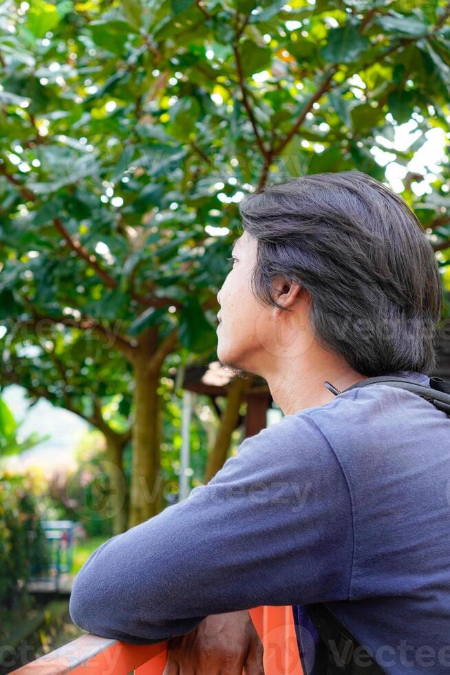 lado Visão do uma homem sentado pensativamente com dele mãos em repouso em uma segurança cerca contra uma pano de fundo do árvores foto