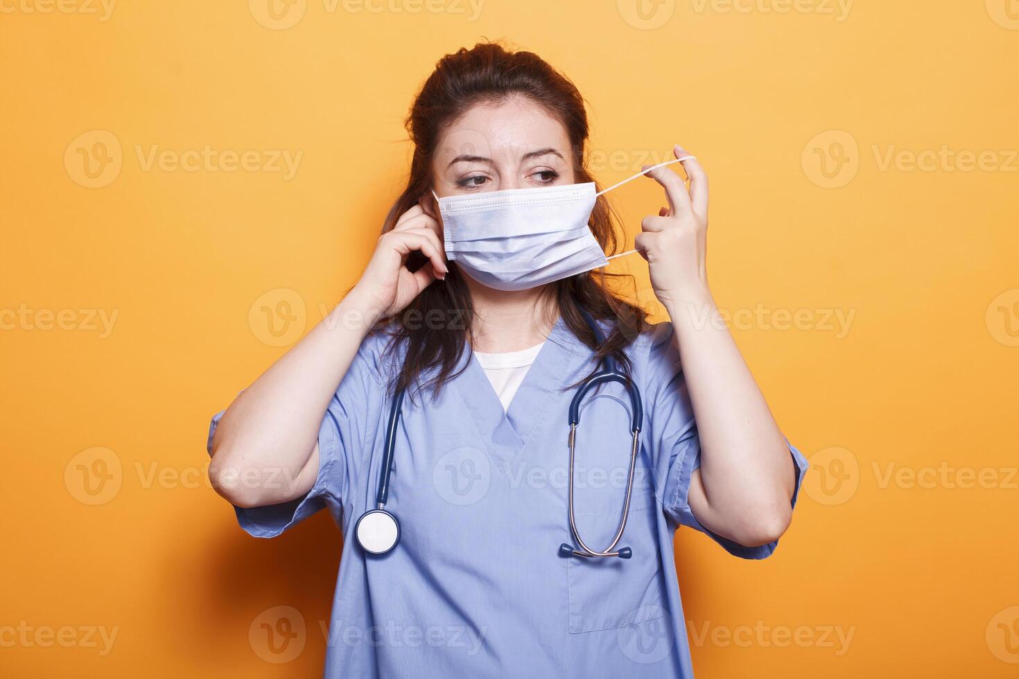 enfermeira praticante colocando em face mascarar para proteção dentro frente do Câmera. fêmea médico médico vestindo esfrega e estetoscópio tendo segurança mascarar contra coronavírus durante pandemia. foto