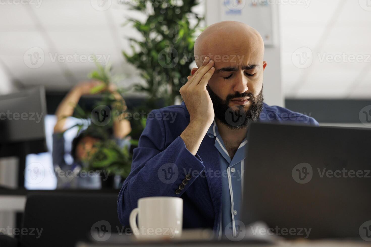 cansado árabe homem de negocios com dor de cabeça trabalhando dentro o negócio escritório. sobrecarregado executivo Gerente sofrimento a partir de enxaqueca e fricção templos enquanto analisando começar acima companhia relatório em computador portátil foto