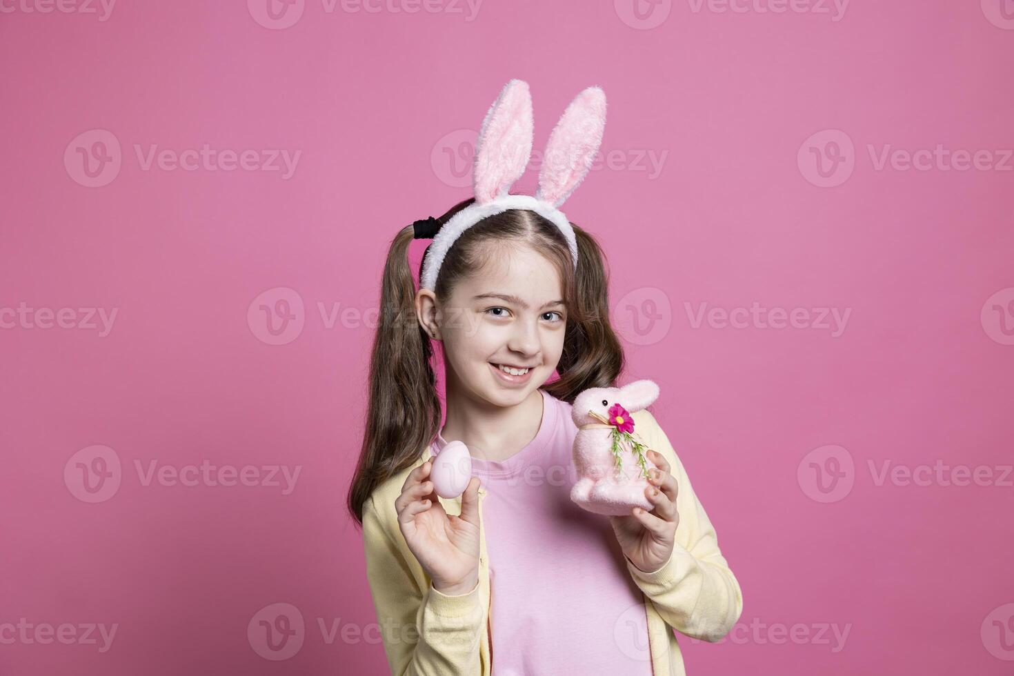 brincalhão jovem menina posando com colori ovos e uma recheado Rosa coelho, vestindo Coelho orelhas e sorridente dentro frente do Câmera. pequeno adorável criança mostrando dela festivo Páscoa decorações e brinquedos. foto