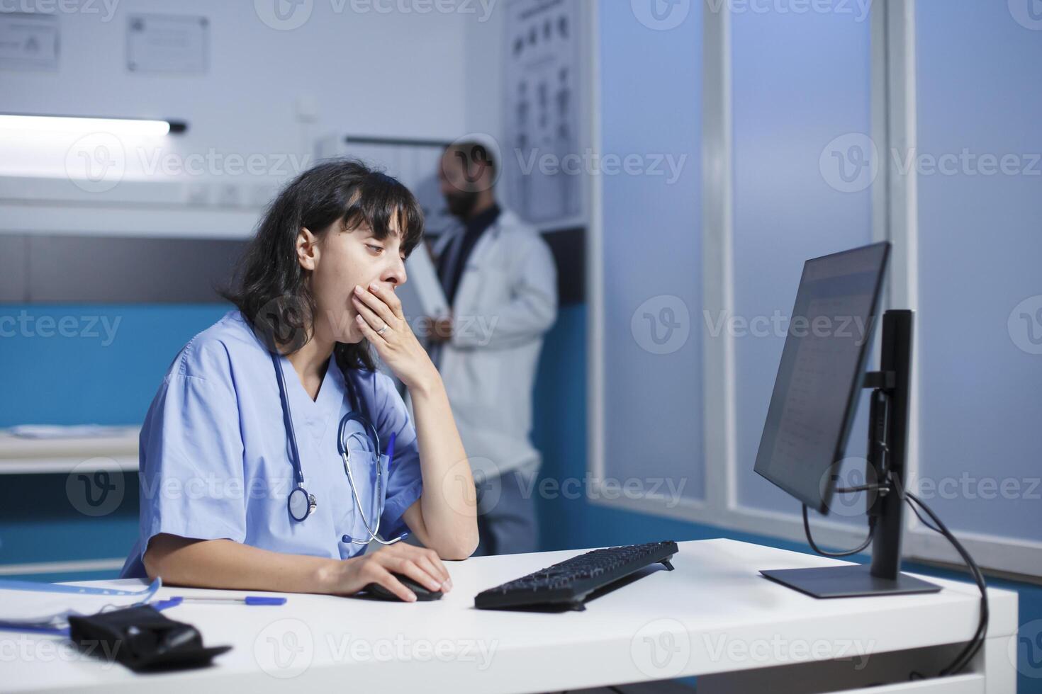 cansado fêmea praticante dentro moderno hospital escritório, usando digital tecnologia para colaboração e comunicação. dedicada enfermeira dentro azul esfrega cansado revendo médico dados em Área de Trabalho computador. foto