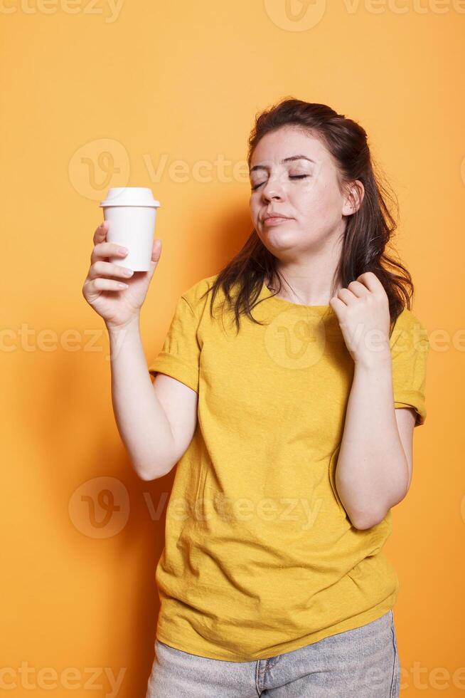 cansado mulher posando contra isolado laranja fundo dentro casual roupas, segurando café xícara, relaxante com fechadas olhos. sonolento expressão do jovem morena senhora retrata exaustão e cansaço. foto