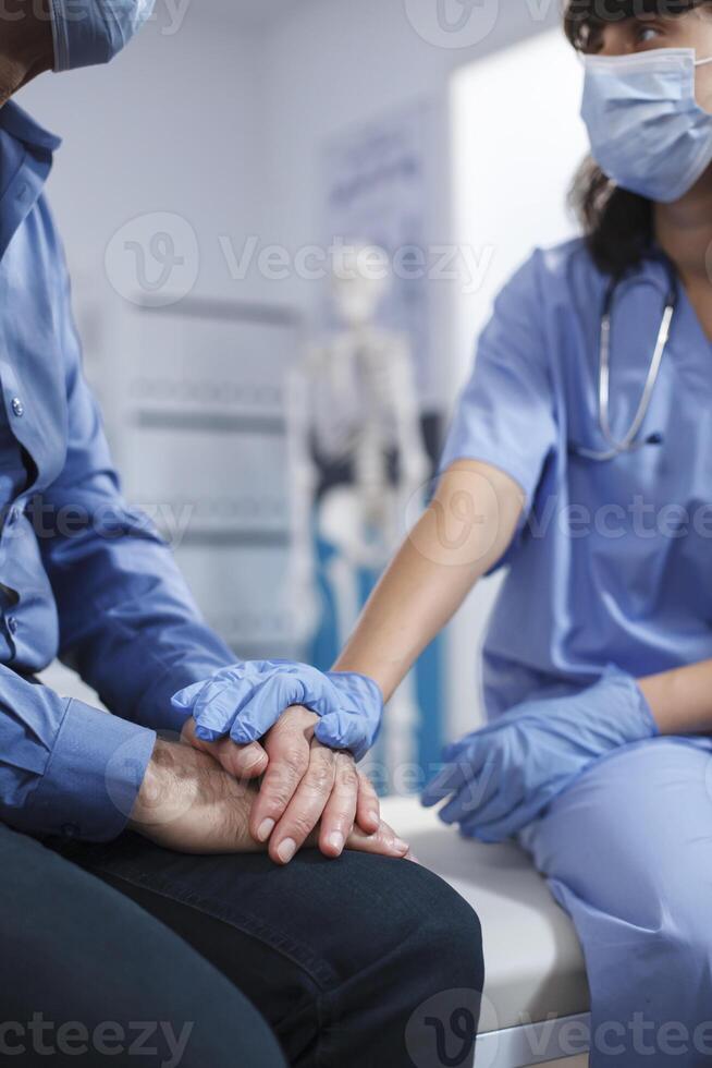 fechar-se do fêmea enfermeira segurando mãos do Senior paciente às hospital escritório. médico praticante vestindo luvas assistências aposentado velho homem depois de uma quiropraxia exame e diagnóstico. foto