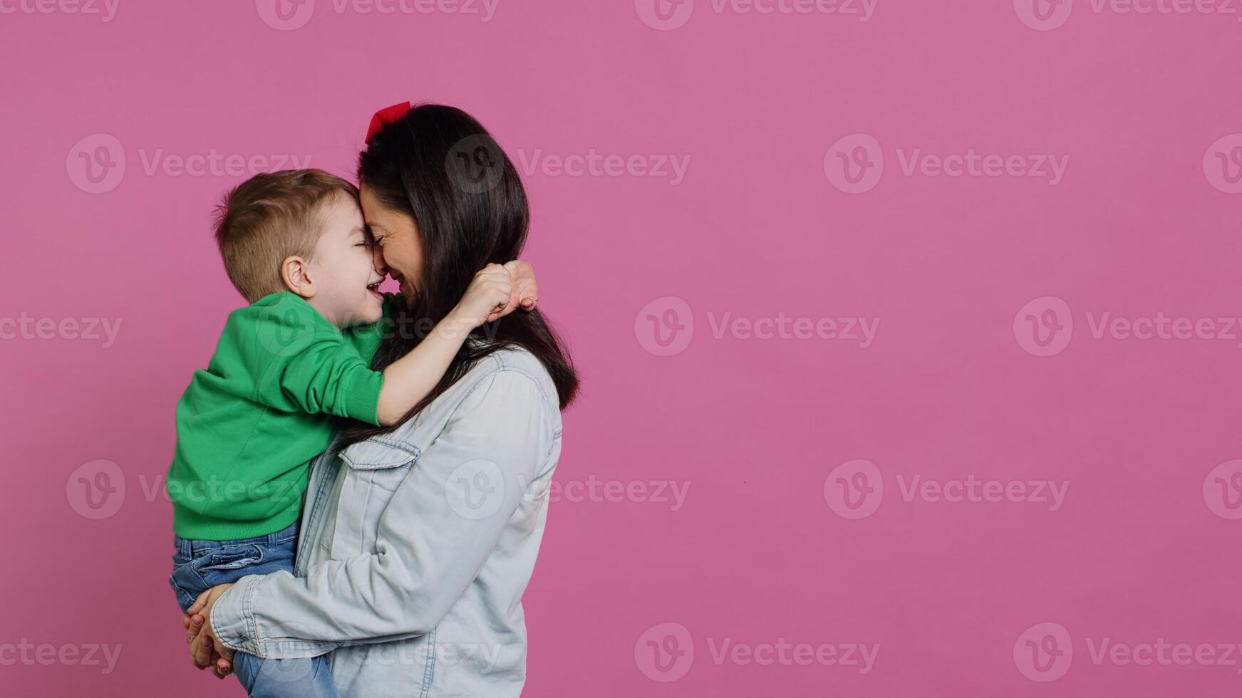 adorável pequeno Garoto abraçando dele mãe e jogando em volta, tendo Diversão contra Rosa fundo. jovem criança pequena posando com dele mãe segurando ele, rindo e ser alegre. Câmera uma. foto