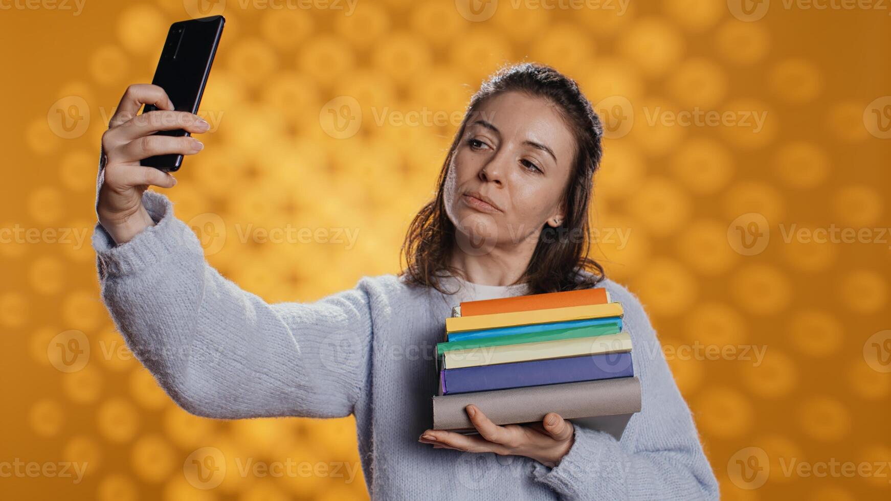 sorridente mulher segurando pilha do livros, desfrutando lendo passatempo, levando selfies com celular. alegre senhora com pilha do romances dentro braços fazendo As fotos com Móvel telefone, estúdio fundo, Câmera b