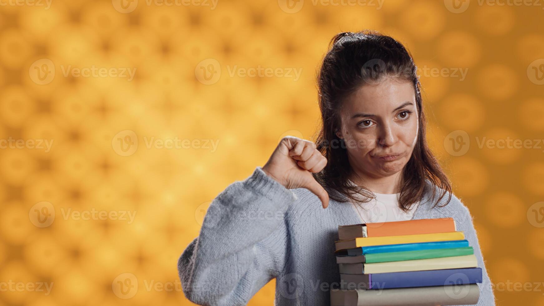 retrato do mulher com fazendo beicinho expressão segurando pilha do livros, mostrando desaprovação do lendo passatempo. aborrecido senhora com pilha do romances fazendo polegares baixa mão gesticulando, estúdio fundo, Câmera uma foto