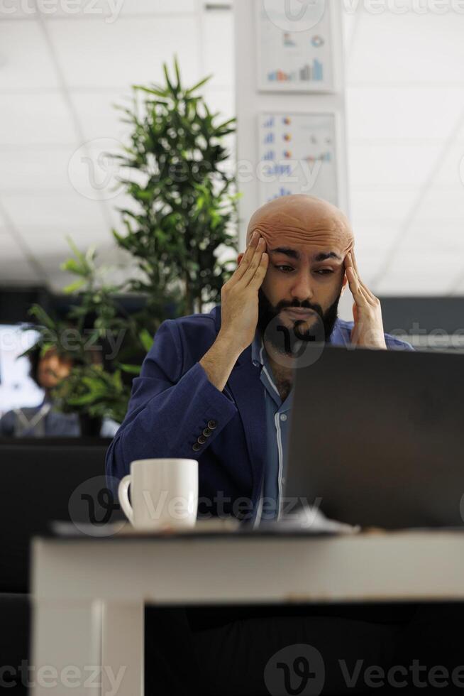 Exausta árabe empreendedor com enxaqueca escrevendo o negócio plano em computador portátil dentro escritório. sobrecarregado empregado tendo dor de cabeça enquanto analisando começar acima companhia marketing estratégia foto