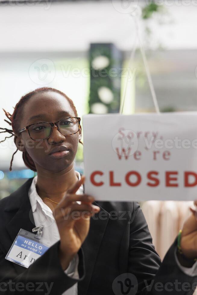 mulher segurando fechadas placa em moda roupas loja frente porta, informando clientes sobre o negócio horas fim. vestuário fazer compras africano americano assistente suspensão placa em viúva foto