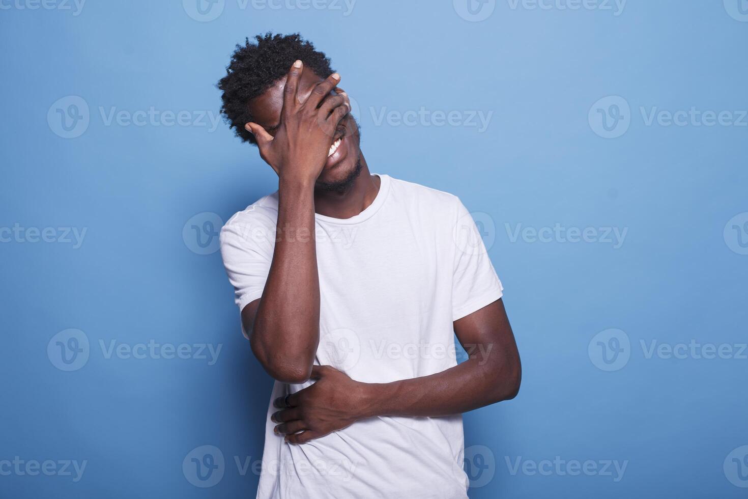 retrato do alegre jovem homem com afro, rindo às Câmera enquanto em pé dentro estúdio com azul fundo. africano americano cara com dele mãos cobertura olhos e estômago é sorridente e rindo. foto