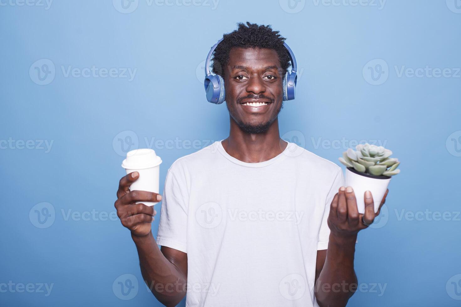 retrato do feliz africano americano homem em pé dentro frente do uma azul fundo com uma em vaso plantar enquanto carregando uma bebida. sorridente Preto cara segurando uma café copo e vestindo sem fio fones de ouvido. foto