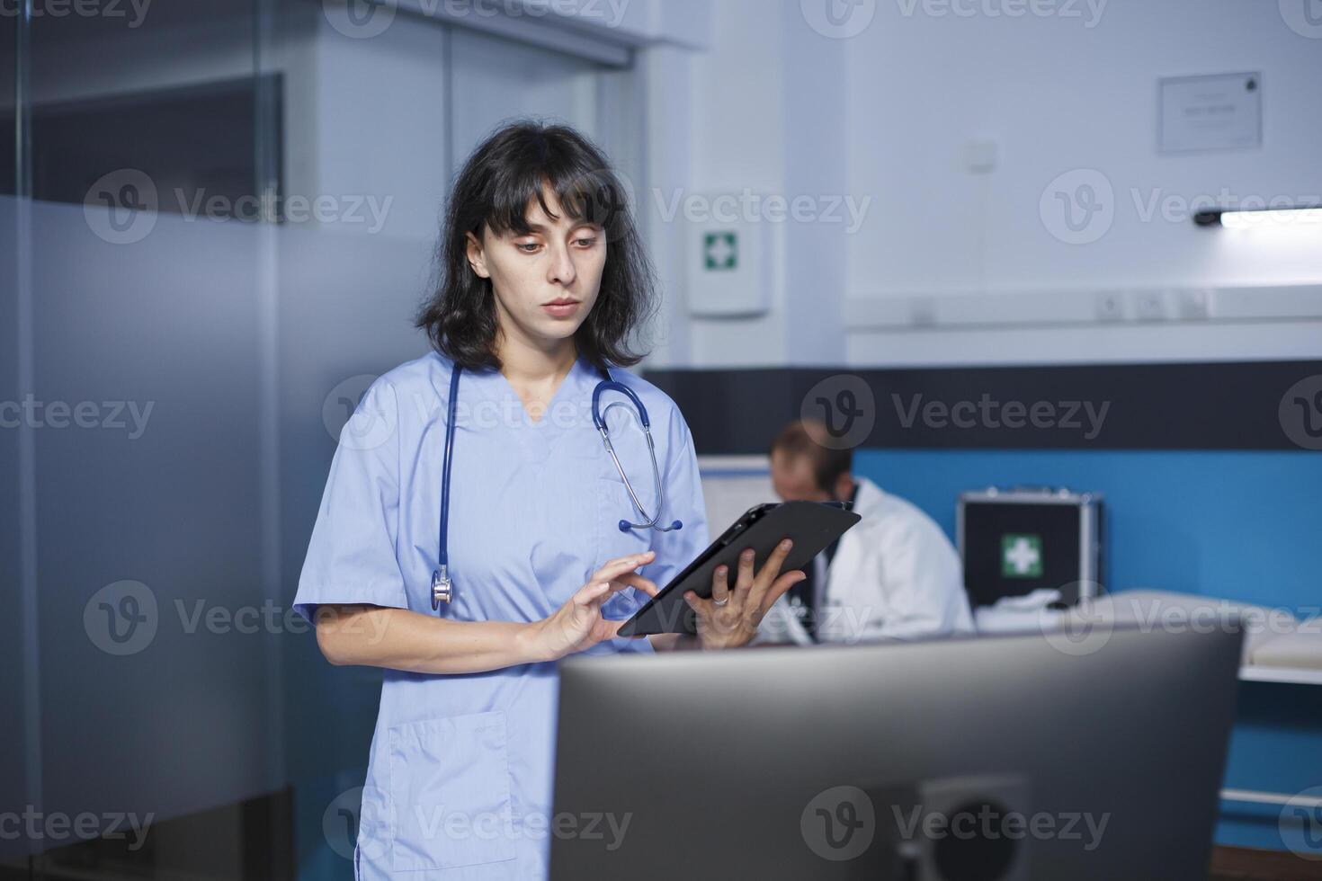 dedicada fêmea enfermeira com estetoscópio comparando médico em formação em dela tábua e Área de Trabalho pc. em pé dentro clínica escritório é caucasiano médico utilizando moderno tecnologia para consultas. foto