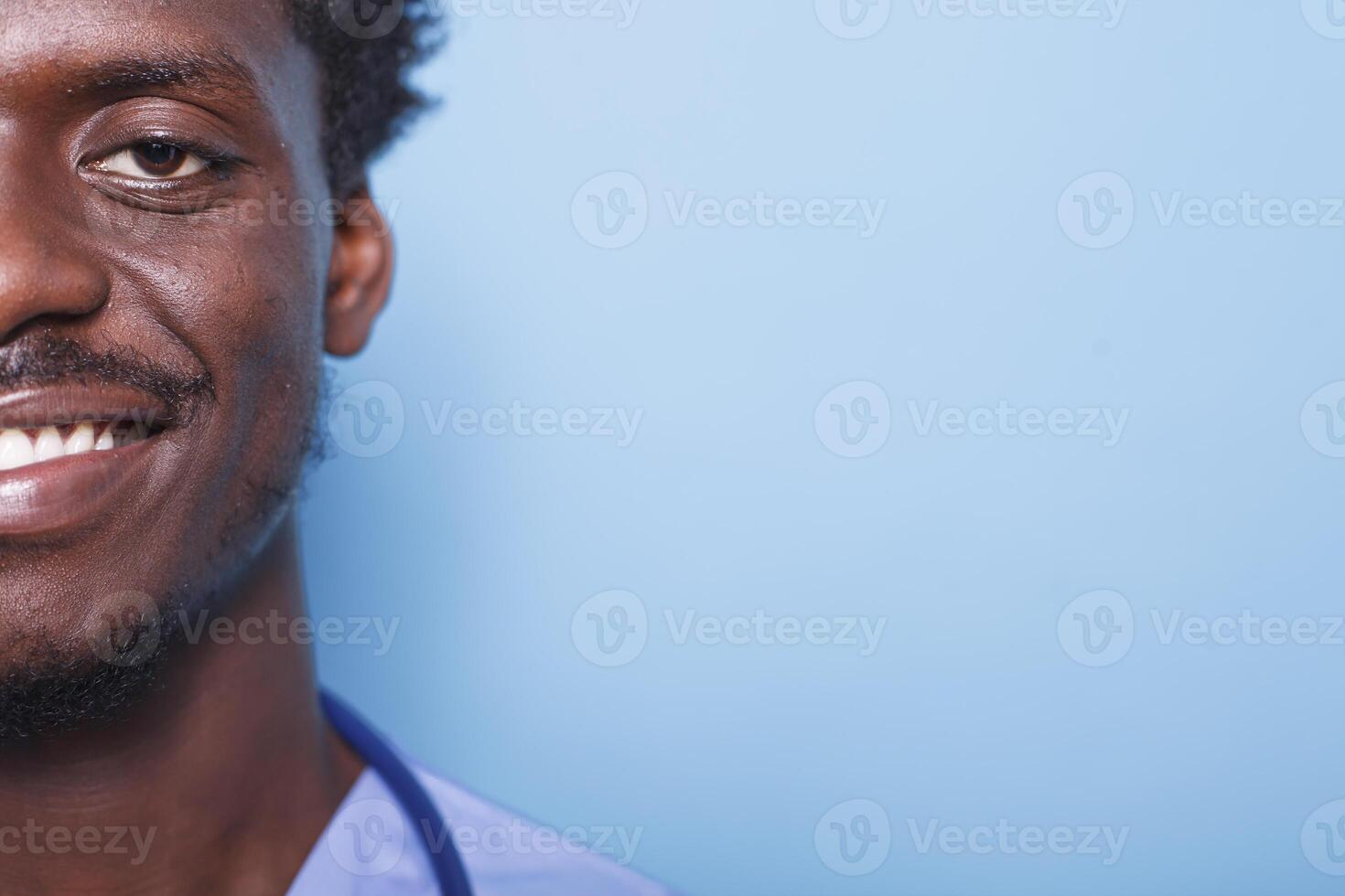 fechar-se retrato do alegre masculino enfermeira vestindo esfrega e estetoscópio em uma azul fundo. metade face, cortada imagem do confiante africano americano médico médico sorridente às Câmera. foto