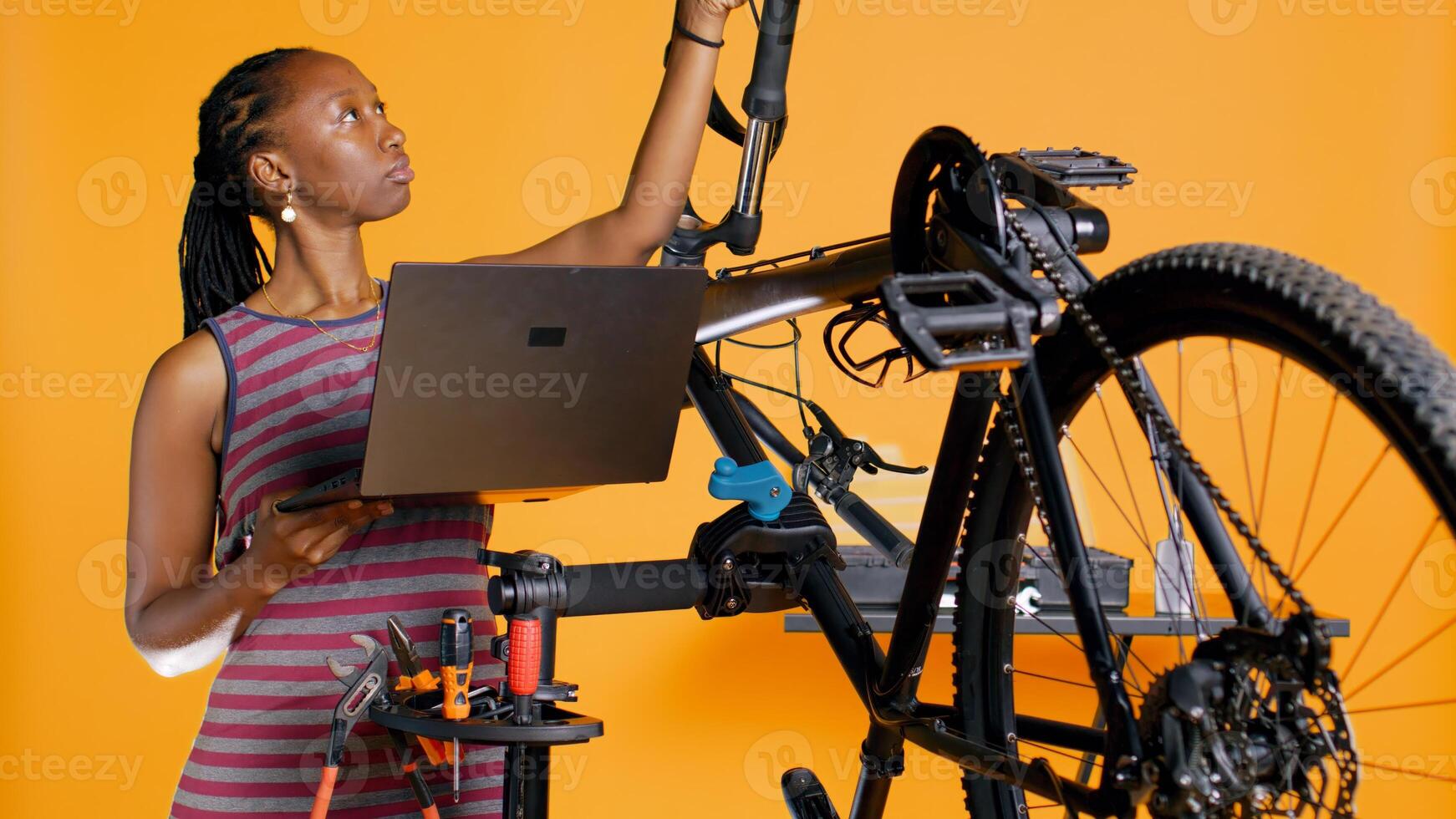 técnico com computador portátil dentro mão analisando quebrado bicicleta sobre estúdio fundo aguardando reparar. especialista usando caderno para Veja para substituição partes para bicicleta em reparar ficar em pé, Câmera b foto