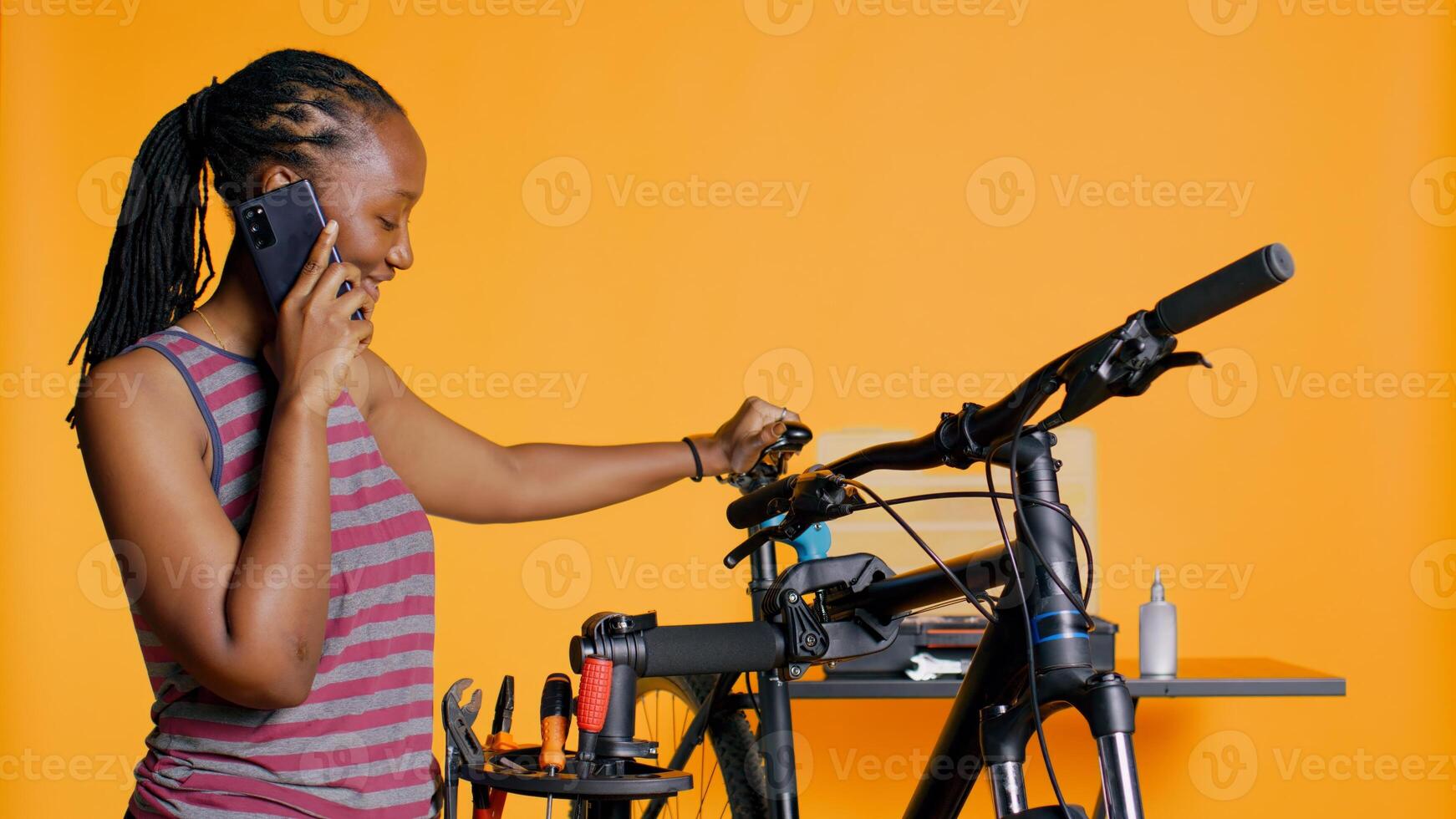 alegre engenheiro às trabalhos levando pausa a partir de reparação bicicleta para conversa em telefone com amigos, pegando acima, estúdio fundo. sorridente mecânico em Smartphone ligar com companheiros enquanto fixação bicicleta, Câmera b foto