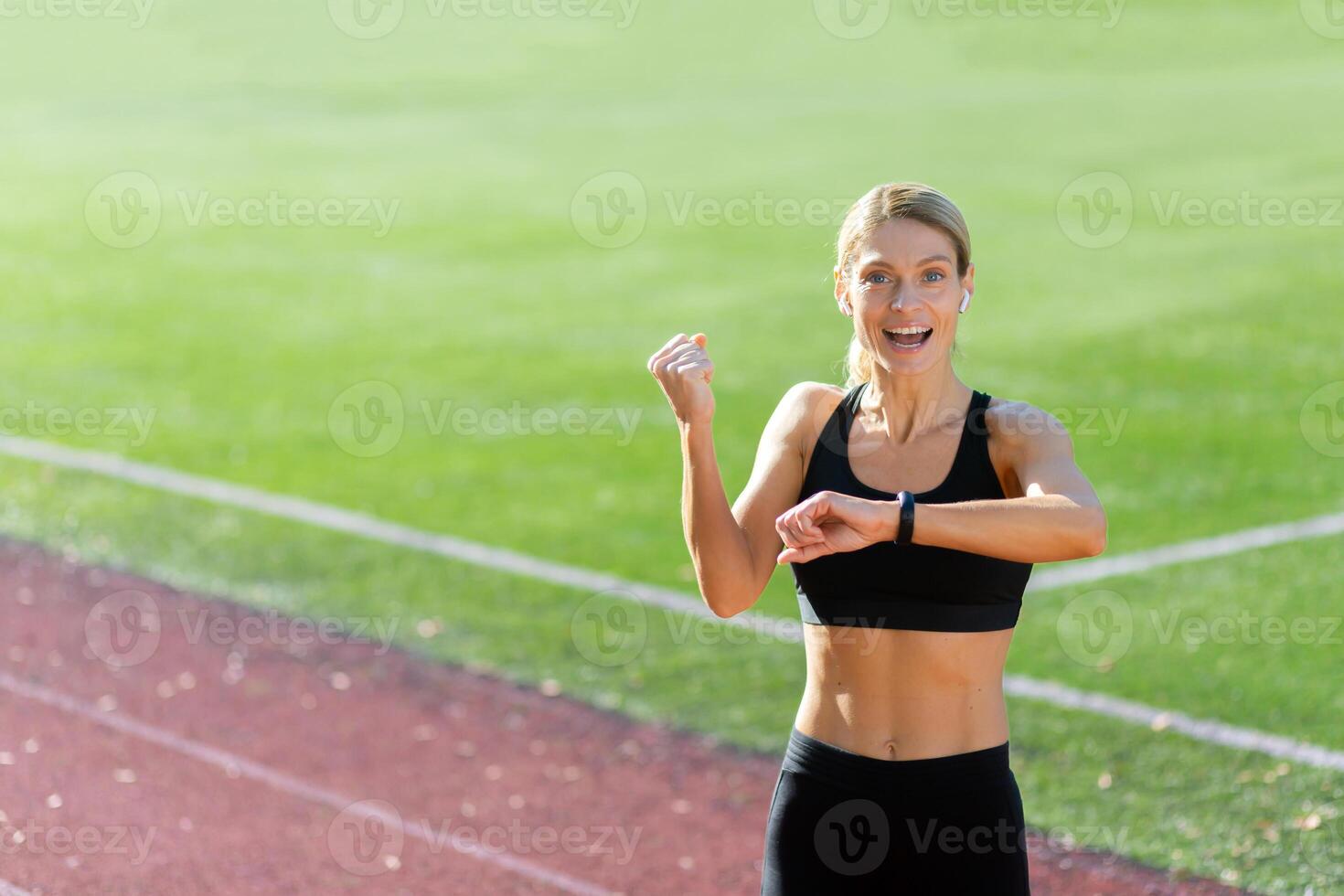 desportivo jovem mulher atleta em pé dentro a estádio vestindo fones de ouvido e sorridente às a Câmera, verificação a Tempo em a ginástica pulseira, mostrando uma vitória gesto com dela mão. foto