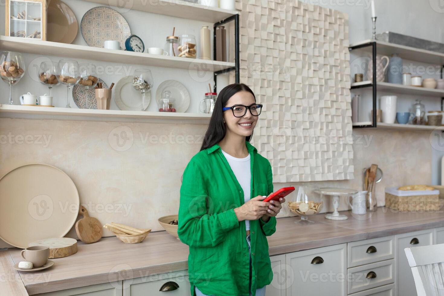 uma jovem lindo mulher dentro óculos e uma verde camisa é em pé dentro a cozinha às casa, digitando uma mensagem em a vermelho telefone, conversando com dela amigos, usando uma Novo aplicativo. ele parece às a Câmera, sorri. foto