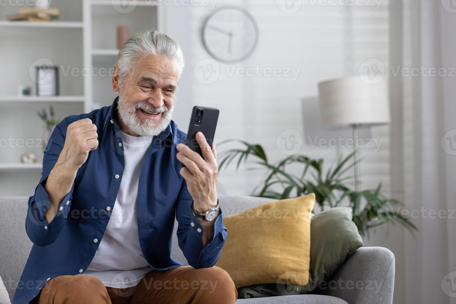 alegre maduro homem com branco cabelo jubilosamente comemora enquanto olhando às dele Smartphone, sentado confortavelmente dentro uma vivo quarto contexto. foto