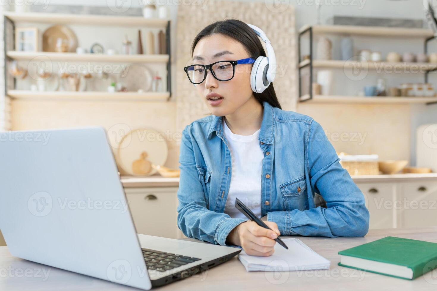 jovem ásia aluna estudando às casa remotamente, mulher assistindo conectados curso sentado dentro cozinha usando computador portátil e bloco de anotações foto