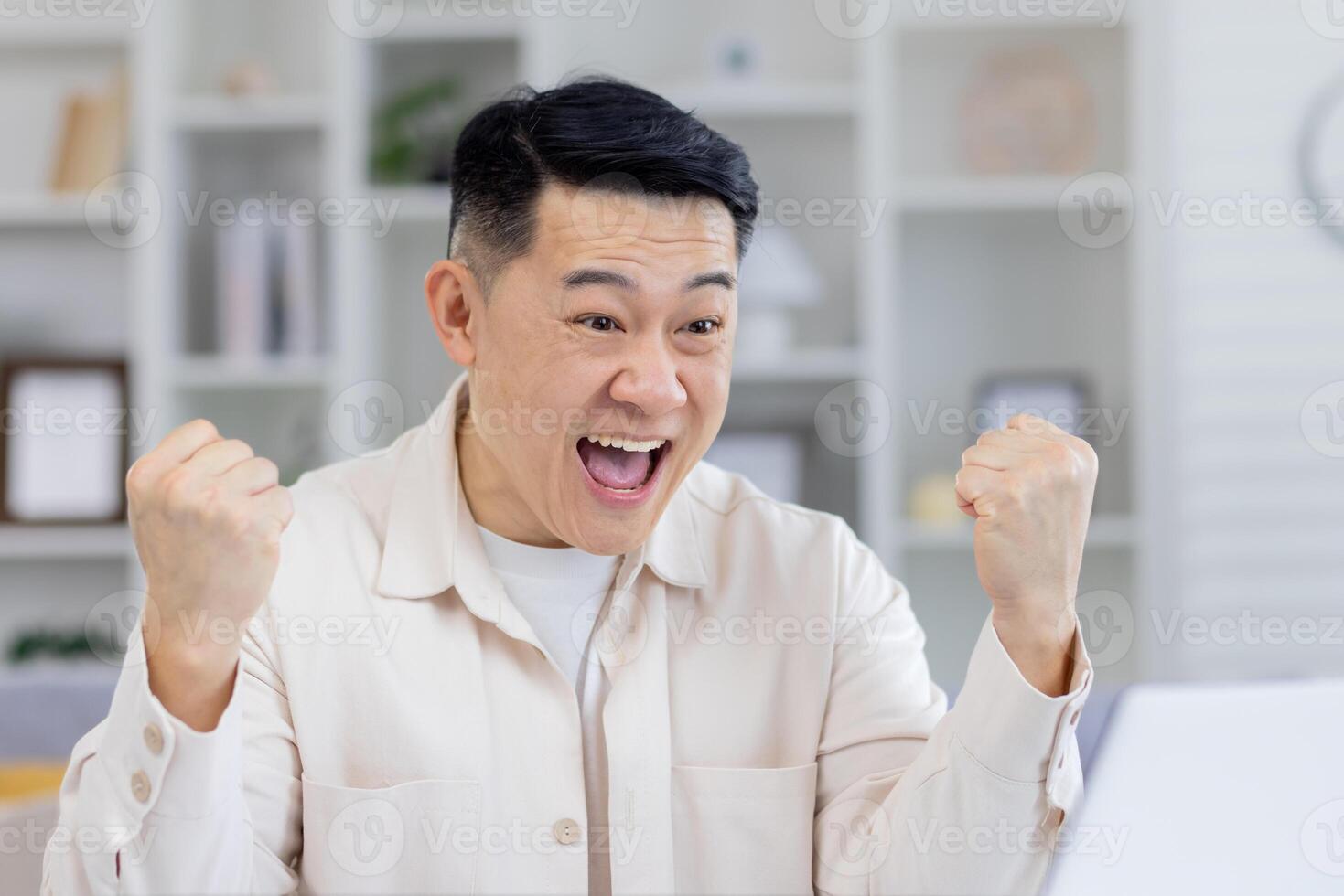 meia idade ásia homem dentro uma creme camisa alegremente a comemorar enquanto olhando às dele computador portátil tela, evidentemente emocionados de Boa notícia ou sucesso às lar. foto