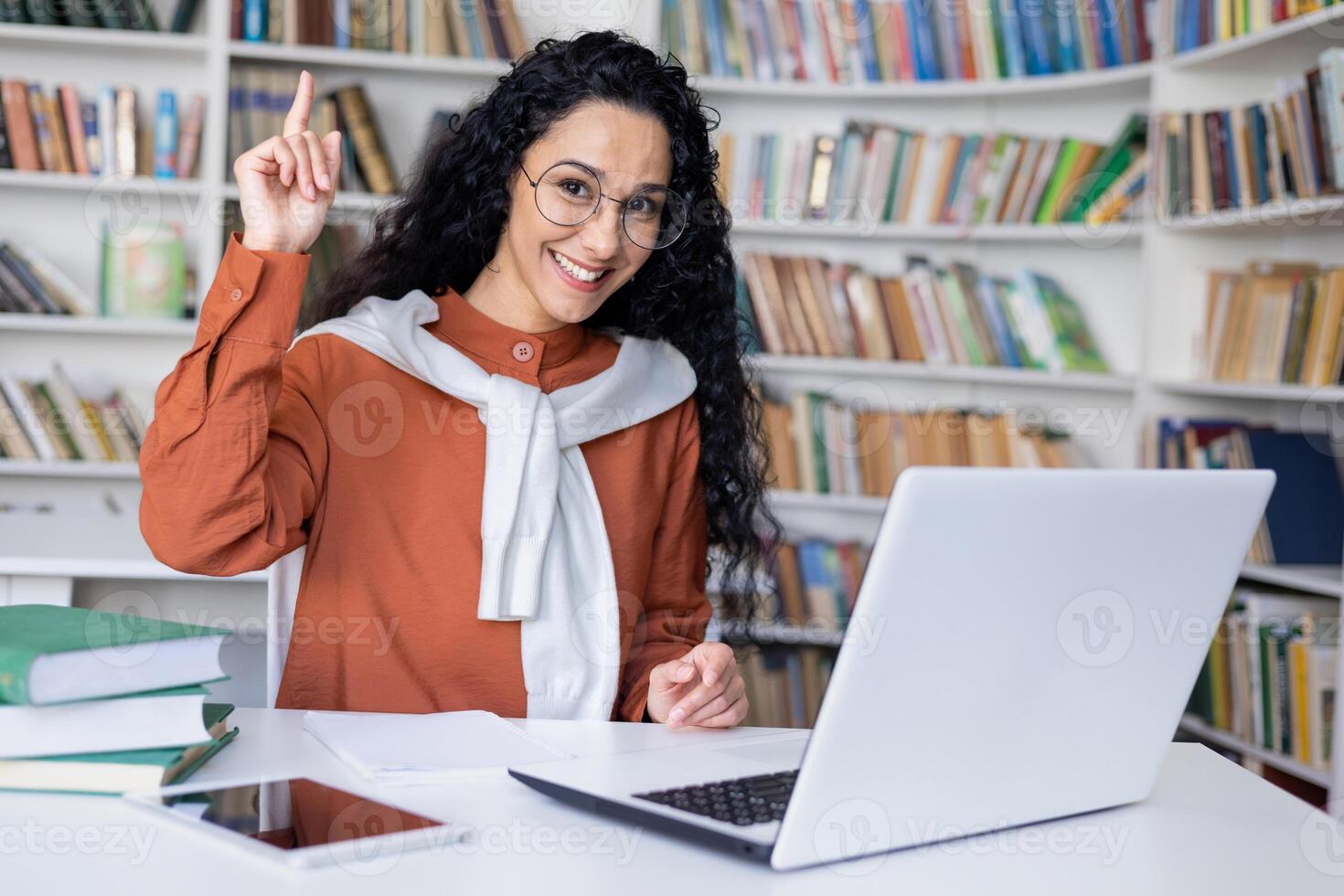 Deleitado universidade aluna fazer eureka gesto enquanto sentado de mesa com pc em fundo do estantes. inteligente fêmea fazendo dever de casa e obtendo certo responda para matemática tarefa. foto