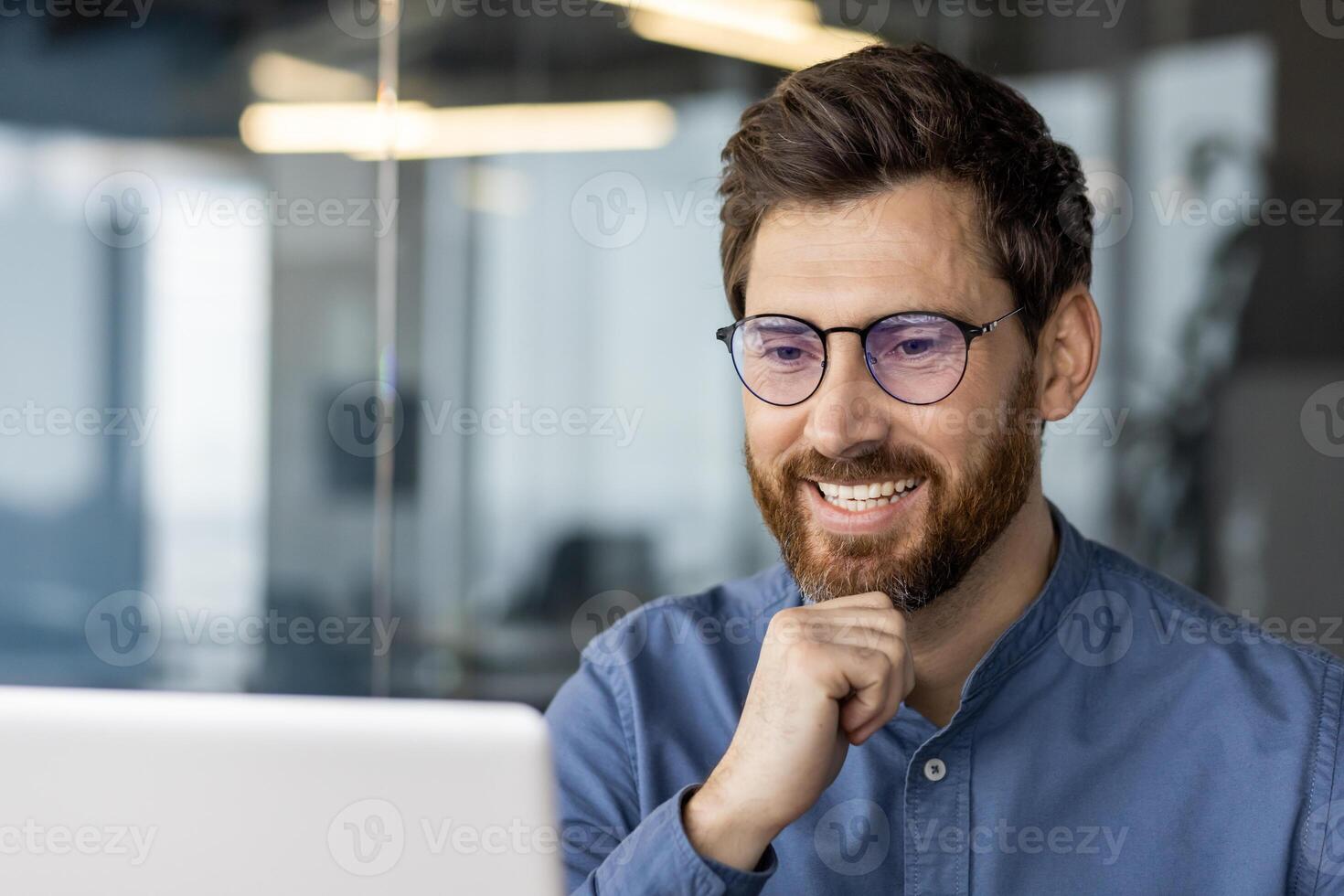 uma conteúdo masculino profissional com uma barba e óculos sorrisos enquanto concentrando em dele computador portátil tela dentro uma moderno escritório contexto. foto