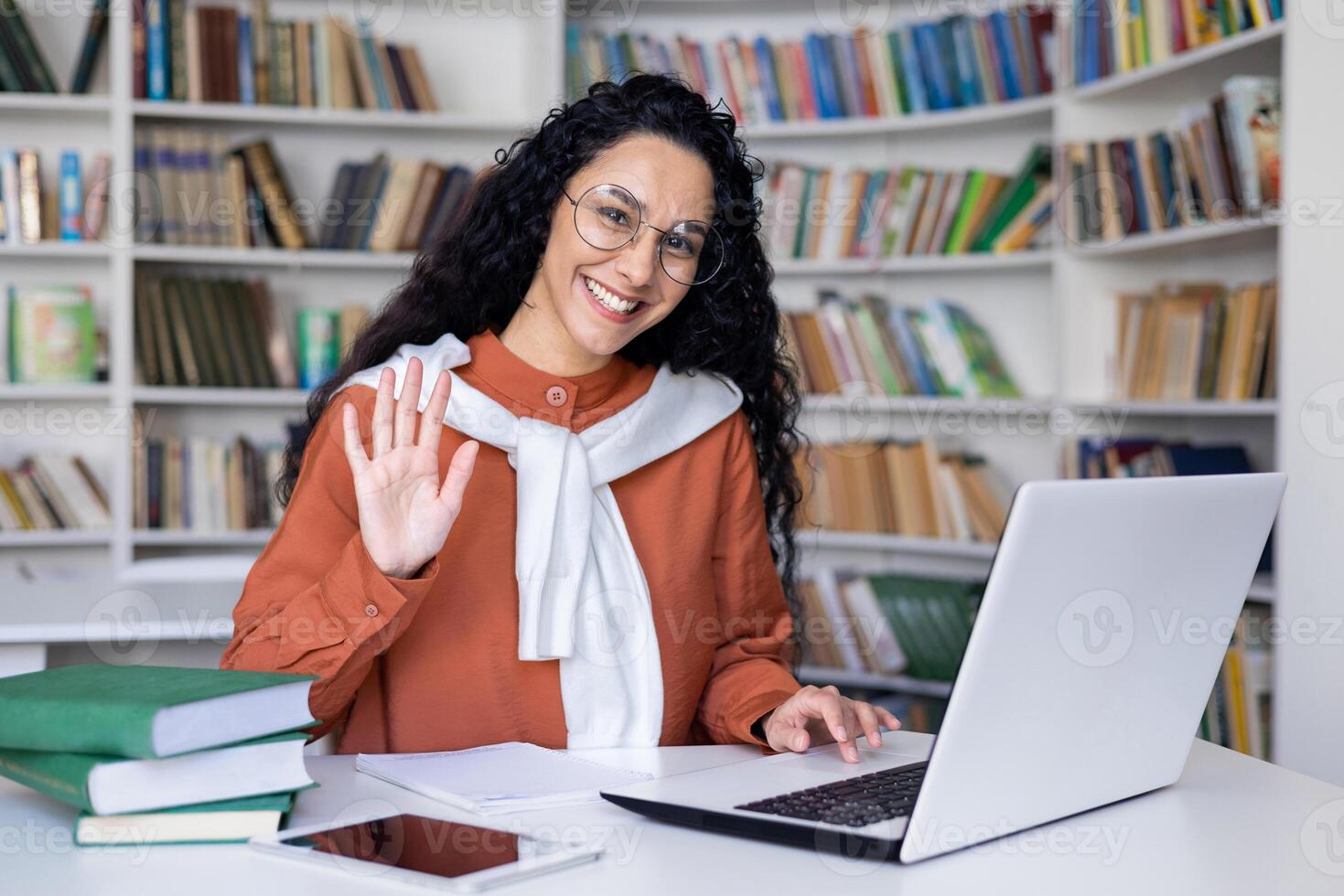 uma jovem fêmea estudante, cercado de livros, ondas às dela computador portátil tela dentro uma biblioteca contexto, sugerindo uma virtual olá. foto