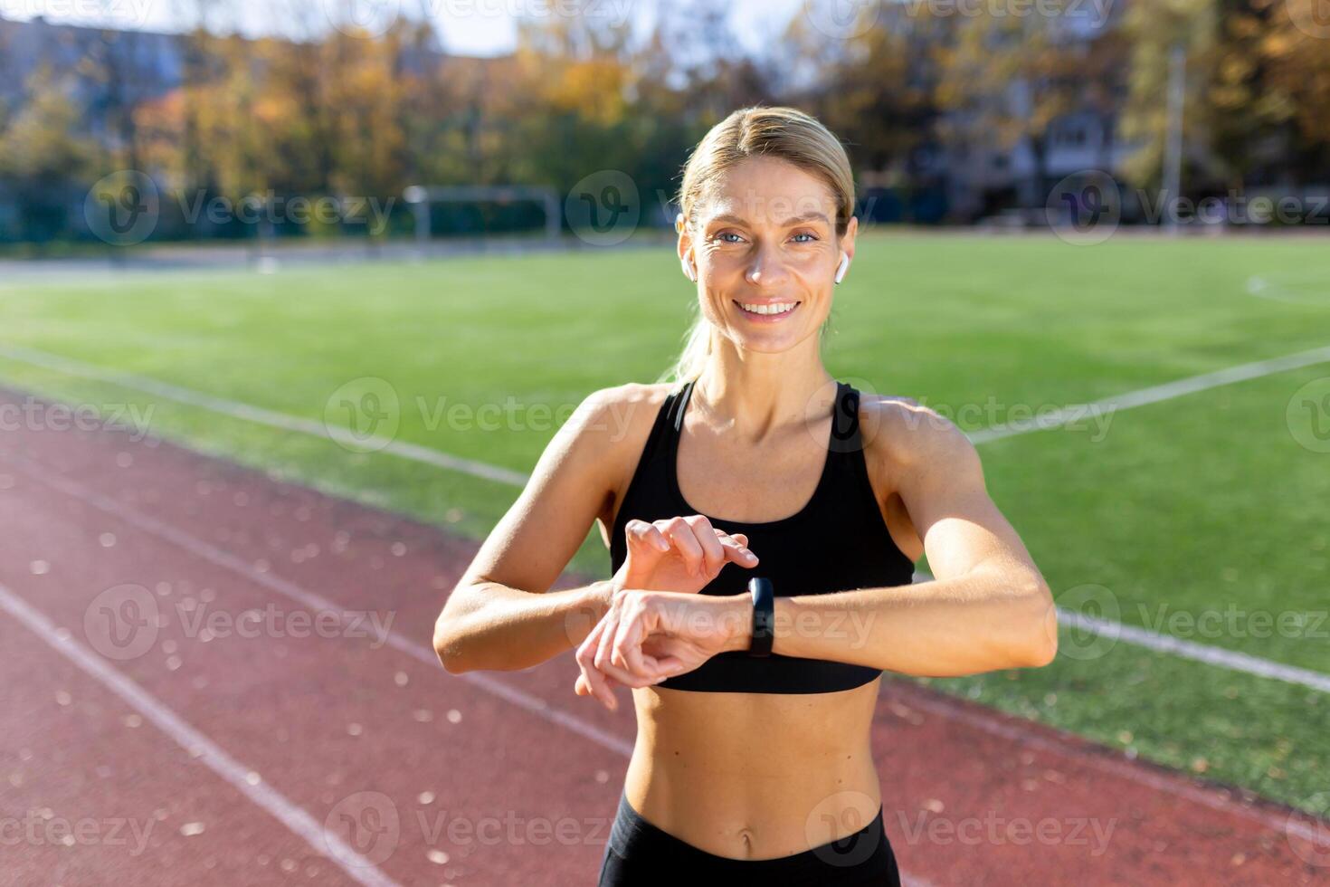 retrato do uma desportivo jovem mulher em pé às a estádio vestindo fones de ouvido e sorridente às a Câmera, verificação a Tempo em uma ginástica pulseira, olhando às a Tempo do correndo, Aulas. foto