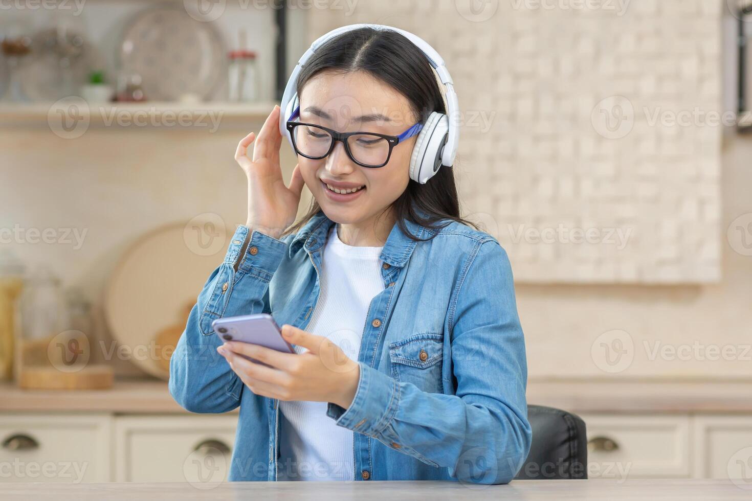 jovem lindo ásia mulher às casa usando fones de ouvido e Móvel telefone para ouço para conectados podcasts, música e audio livros, fechar-se mulher dentro óculos dentro cozinha sorridente e feliz foto