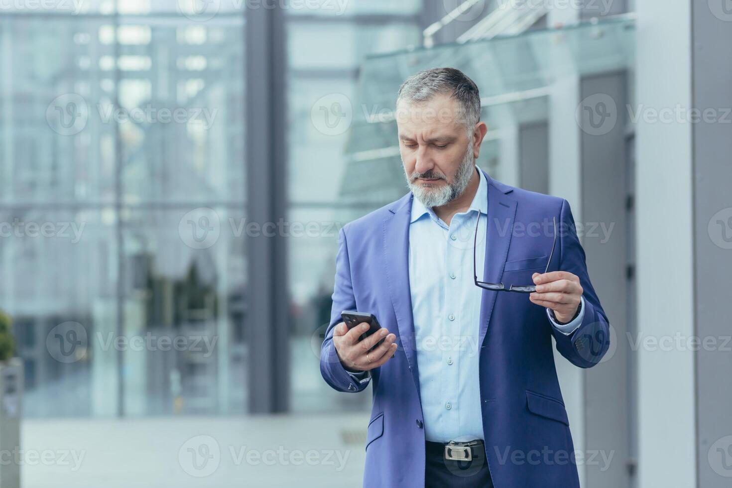 Senior bonito grisalho homem dentro uma terno segurando uma telefone e óculos. concentradamente lê a notícia em a telefone, parece às mensagens. isto é localizado perto uma moderno escritório Centro. foto