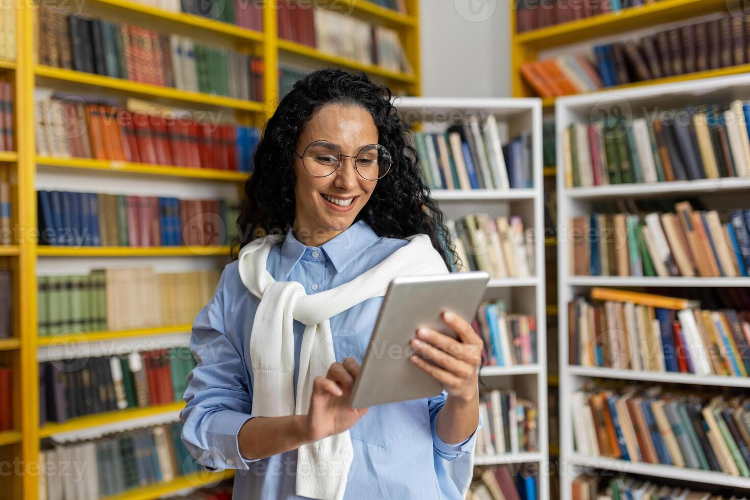 sorridente jovem mulher dentro óculos usando uma digital tábua entre estantes de livros dentro uma biblioteca, representando moderno Aprendendo e tecnologia dentro Educação. foto