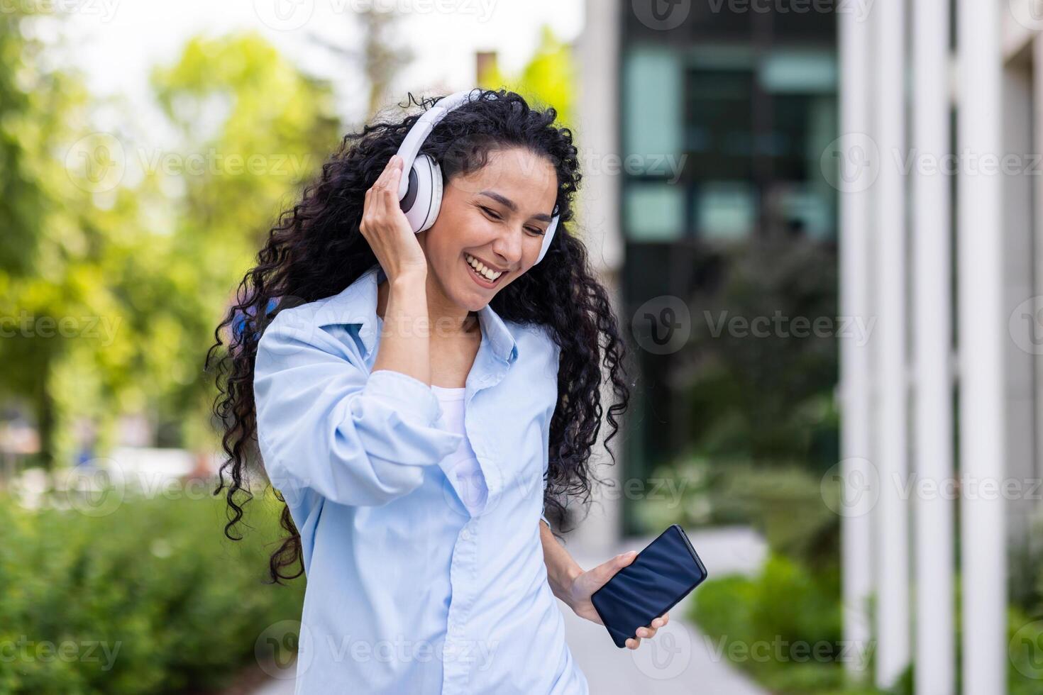 uma alegre, cabelo encaracolado mulher é apanhado dentro uma momento do alegria enquanto ouvindo para música em dela branco fones de ouvido, segurando uma Smartphone, ao ar livre com vegetação e uma construção dentro a fundo. foto