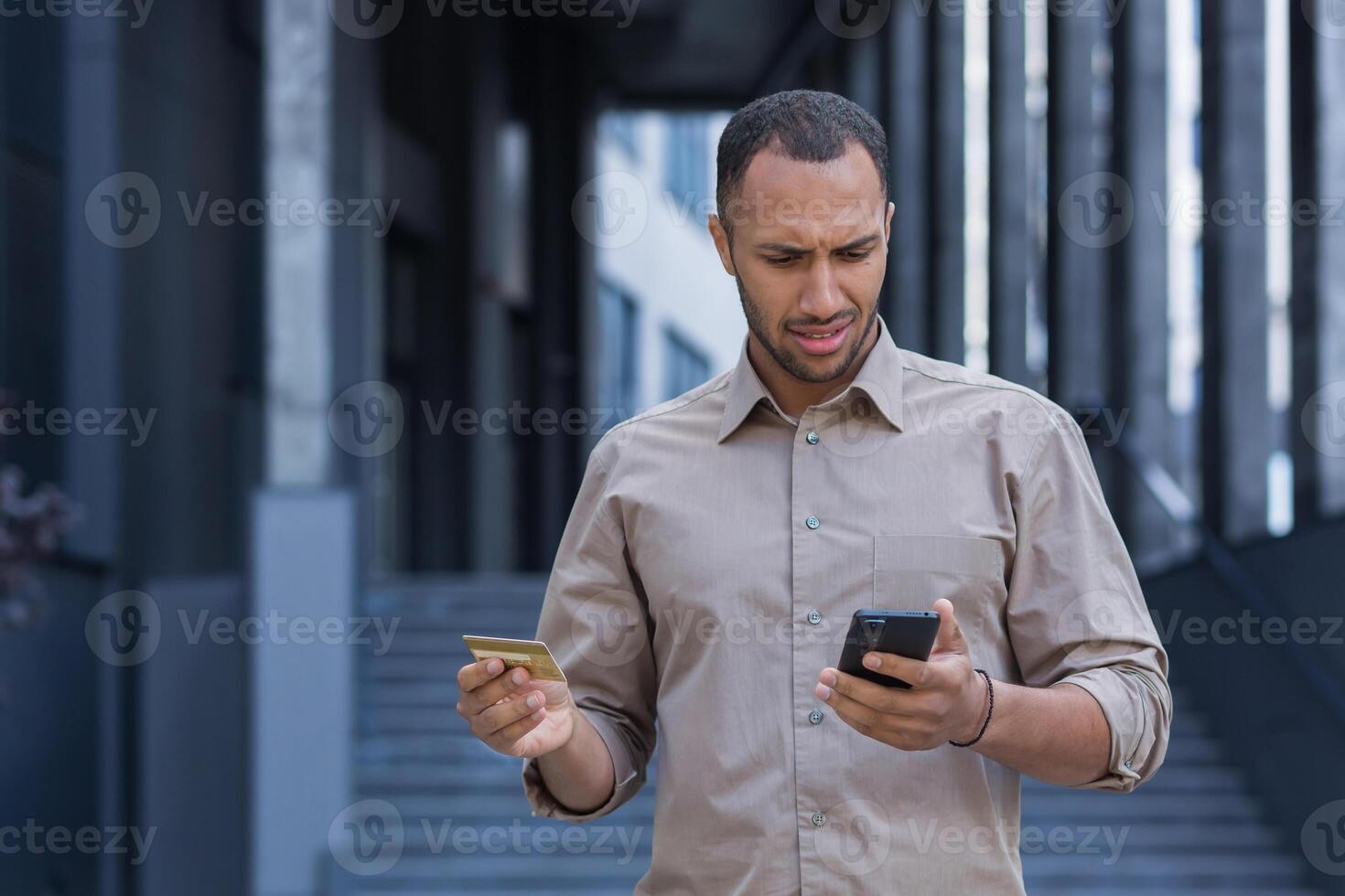 frustrado e chateado africano americano homem lado de fora escritório construção tentando para faço uma compra dentro a conectados loja, homem usando Smartphone e banco crédito cartão, para transferir dinheiro e Comprar conectados foto