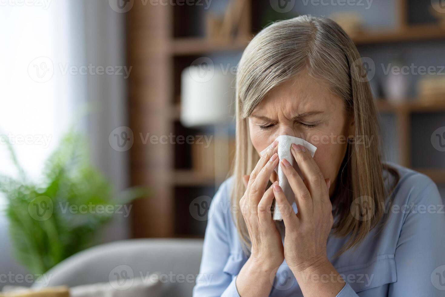 uma maduro mulher experimentando frio sintomas, espirros para dentro uma lenço de papel enquanto sentado dentro uma acolhedor vivo sala. saúde e bem estar conceito. foto